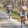 A group of runners participating in an outdoor race on a gravel path surrounded by pine trees. The foreground features a woman in a white tank top and blue sneakers leading ahead of a man wearing a 'Protect This House' shirt with race bib number 9. Additional runners are visible further back on the trail.