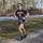 A male runner wearing bib number 685 is competing in an outdoor road race along a gravel and paved path surrounded by tall pine trees. He is wearing a humorous black t-shirt reading 'Exercise? I thought you said Extra Fries' with a graphic of french fries. The setting appears to be a wooded park or trail race course during late fall or winter based on the bare trees.
