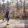 A bald man is running at speed along a gravel path through a wooded area. The image uses panning photography technique, keeping the subject relatively sharp while the background is heavily motion-blurred to convey a sense of speed. He is dressed in athletic wear and appears to be in mid-stride during an outdoor run.