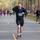 A road race is taking place on a tree-lined paved road, likely in a park or forested area. The foreground runner, bib number 696, is drinking from a small cup while running, a common sight at hydration stations during races. Numerous other runners are visible in the background, spread across the road.
