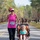 A woman wearing a pink tank top and race bib number 221 is running in an organized road race while pushing a young child in an orange and black jogging stroller. The event appears to take place on a closed road lined with trees, with a large group of runners visible in the background. The setting suggests a military base race event, indicated by the bib text referencing 'MCAS Cherry Point, NC'.