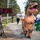 A fun run or charity race event taking place on a gravel path lined with pine trees. Two participants are wearing inflatable T-Rex dinosaur costumes — one large orange and one smaller green — among other runners in casual athletic wear. A digital race clock on a tripod displays the time 46:58.