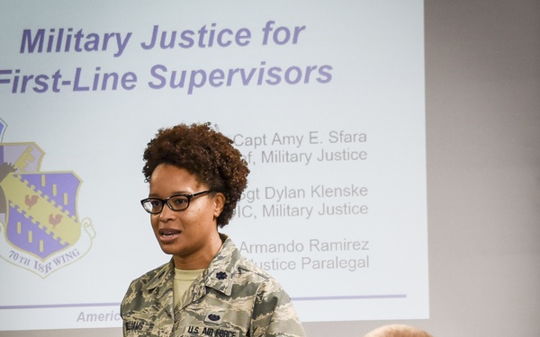 A U.S. Air Force officer is presenting a briefing titled 'Military Justice for First-Line Supervisors' to a group of uniformed military personnel. The presenter is wearing the Airman Battle Uniform (ABU) with U.S. Air Force insignia and appears to be addressing an audience seated in front of her. The presentation slide visible in the background lists presenters including Capt Amy E. Sfara and references the 70th ISR Wing.