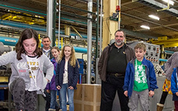 A young girl is stomping on a pink air-pressure pad connected via a blue tube to a small rocket launcher on an industrial facility floor. A group of children and adults stand watching in the background, suggesting this is a demonstration or educational event. The setting appears to be a manufacturing plant or warehouse during what looks like a family or school tour.
