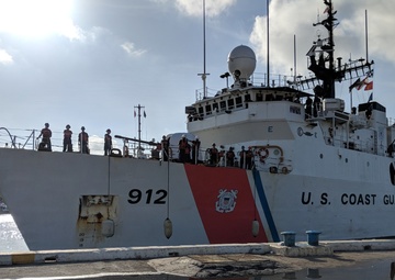 A U.S. Coast Guard cutter numbered 912 is docked at a pier, with multiple crew members visible on deck. The vessel displays the distinctive USCG livery featuring a red and blue diagonal racing stripe and the Coast Guard emblem. The ship's reflection is clearly visible in standing water on the dock surface.
