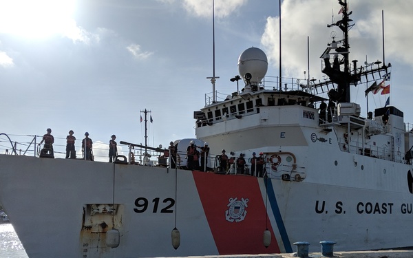 A U.S. Coast Guard cutter numbered 912 is docked at a pier, with multiple crew members visible on deck. The vessel displays the distinctive USCG livery featuring a red and blue diagonal racing stripe and the Coast Guard emblem. The ship's reflection is clearly visible in standing water on the dock surface.