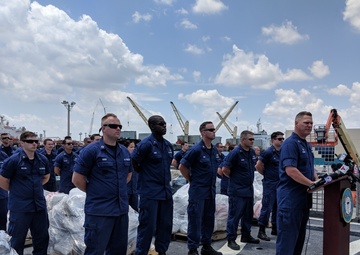 A formal press conference is taking place on the deck of a U.S. Coast Guard vessel at a port facility. A senior officer is speaking at a podium equipped with multiple microphones, while a large group of uniformed Coast Guard personnel stands at attention behind him. Pallets of seized contraband, wrapped in plastic, are visible on the deck, suggesting this is a drug interdiction offload ceremony.