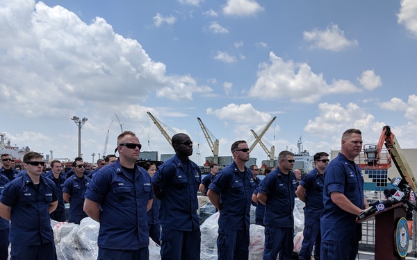 A formal press conference is taking place on the deck of a U.S. Coast Guard vessel at a port facility. A senior officer is speaking at a podium equipped with multiple microphones, while a large group of uniformed Coast Guard personnel stands at attention behind him. Pallets of seized contraband, wrapped in plastic, are visible on the deck, suggesting this is a drug interdiction offload ceremony.