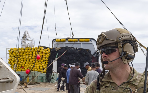 A heavily equipped military or law enforcement operator in multicam camouflage stands guard on the deck of a vessel, holding a radio and wearing tactical gear including a helmet, ear protection, and body armor. In the background, several civilians or detainees are gathered near a large cargo load consisting of yellow buoys and dark netting material. The scene appears consistent with a maritime interdiction or boarding operation at sea.