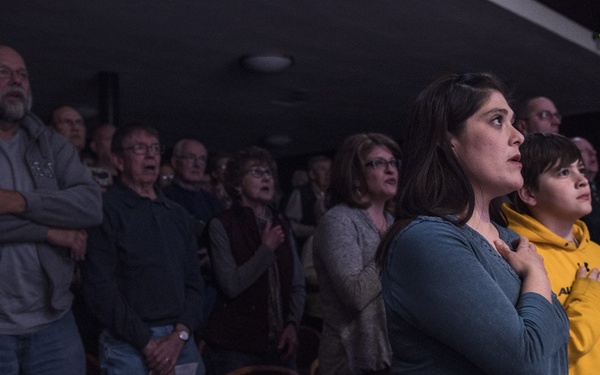 A group of people are standing in an auditorium or theater with burgundy/maroon upholstered seats, with many placing their right hands over their hearts. The gesture and posture of the attendees strongly suggest they are reciting the Pledge of Allegiance or participating in a national anthem. The foreground features a woman in a teal top and a youth in a yellow hoodie, with a crowd of mixed-age adults visible behind them.