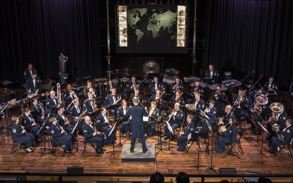 A large military band, dressed in dark blue uniforms, performs on a concert stage before a seated audience. A conductor stands at the center front of the ensemble, facing the musicians. A world map projection is displayed on a screen at the back of the stage.