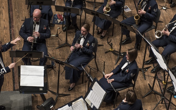 An aerial view of a military band performing in concert, with musicians dressed in dark navy blue uniforms seated around music stands on a hardwood floor stage. A conductor stands on a raised podium at the left, directing the ensemble with a baton. The band includes woodwind and brass instrument players arranged in a semi-circular formation.