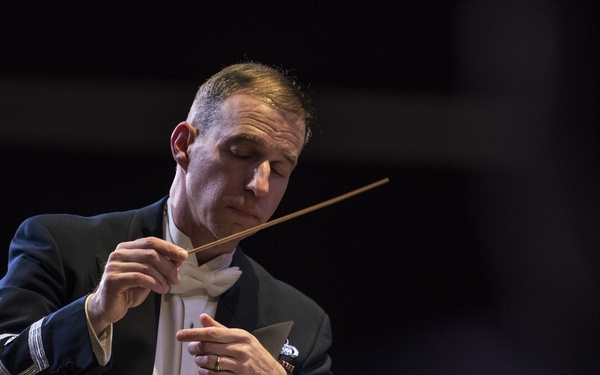 A male orchestra conductor is captured mid-performance, holding a baton in his right hand while gesturing with his left. He is dressed in a formal dark navy military-style dress uniform with silver sleeve insignia, white bow tie, and decorative buttons. The image is dramatically lit against a dark background, emphasizing the conductor's focused expression and precise hand movements.