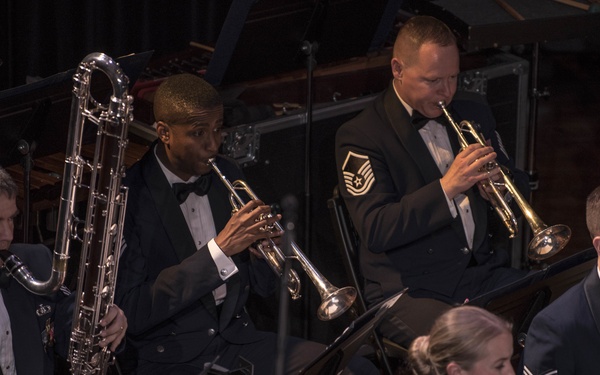 A military band ensemble is performing at a formal concert, with musicians dressed in dark blue dress uniforms with bow ties. The group includes brass and woodwind players, with visible rank insignia identifying them as U.S. Air Force personnel. The performance appears to be taking place on a concert stage under professional lighting.