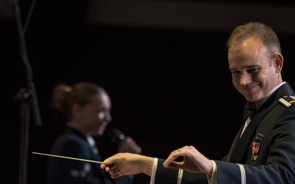 A military officer in dress uniform is conducting an orchestra or band, holding a conductor's baton in his right hand while smiling. In the background, a female military personnel member appears to be speaking into a microphone. Multiple musicians are visible in the foreground, seated with music stands and sheet music.