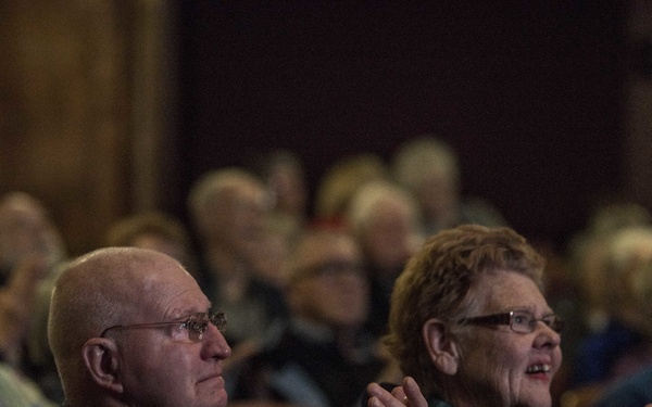 Two elderly individuals are seated in a theater or auditorium, visibly applauding a performance. The foreground shows a bald man wearing glasses and a dark jacket alongside a woman with reddish hair, glasses, and a teal scarf, both clapping enthusiastically. The background reveals a filled audience of similarly aged attendees seated in red upholstered theater chairs.