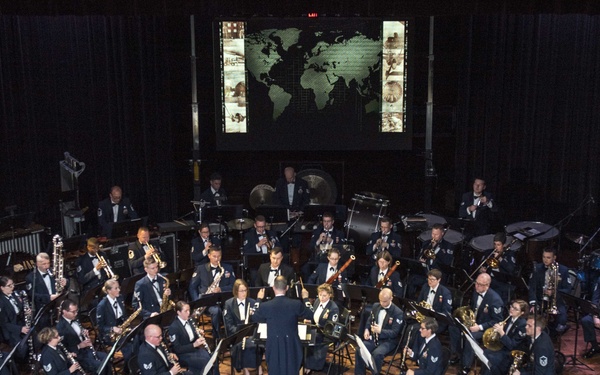 A large military band performs on a formal concert stage, with musicians arranged in a semicircle around a central conductor standing on a podium. All performers are dressed in dark navy blue military dress uniforms. A large projection screen displaying a world map is visible in the background, suggesting a themed or ceremonial performance.