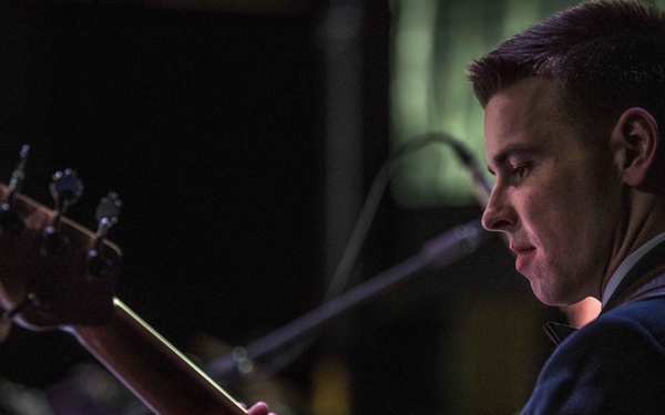A young male U.S. Air Force enlisted member in dress uniform plays a bass guitar during what appears to be a formal military band performance. He is reading from sheet music on a stand while focused on his instrument. The image is a close-up, dramatically lit portrait-style photograph taken in a performance venue.