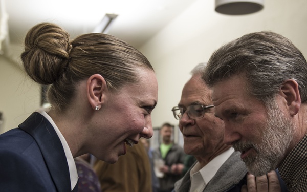 A young woman in a U.S. Air Force uniform with an Airman First Class rank insignia is laughing and conversing with two older men in a crowded indoor setting. The interaction appears warm and celebratory, possibly following a military ceremony or graduation event. Several other people are visible in the background engaged in conversation.