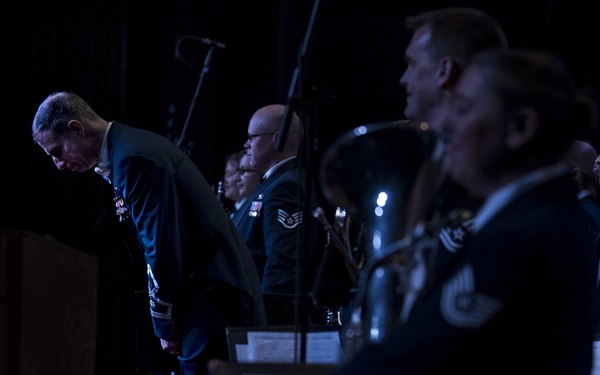 A military band performance on a darkened stage, with uniformed personnel standing at attention or in formation. A senior officer on the left appears to be bowing, likely acknowledging applause at the conclusion of a performance. The scene suggests a formal military concert or ceremonial event.