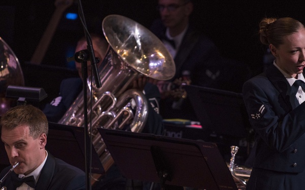 Two U.S. Air Force personnel are performing on stage during a formal military band concert. A male Master Sergeant plays a French horn while seated at a music stand, and a female Technical Sergeant stands at a microphone singing. Both are dressed in formal Air Force dress blue uniforms with visible rank insignia and service ribbons.