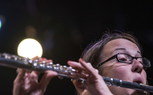A woman wearing black-rimmed glasses and a dark formal jacket with a bow tie is playing a silver transverse flute. She is captured mid-performance under stage lighting against a dark background. The close-up angle emphasizes her focused expression and finger positioning on the instrument.