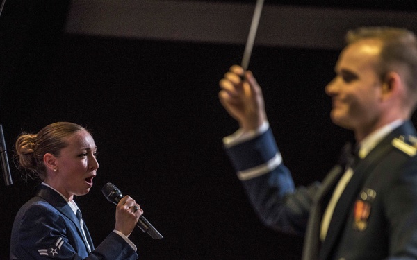 Two U.S. Air Force personnel are performing on stage during a military band concert. A female airman in dress blues is singing into a handheld microphone while a male officer conducts with a baton. Additional band members and audience members are visible in the background.