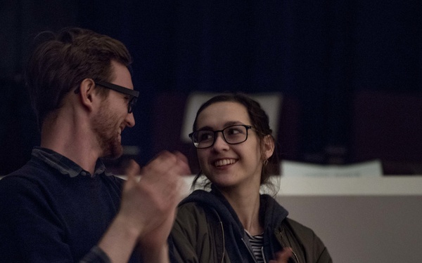 Two people are standing together in a dimly lit indoor setting, both clapping their hands and smiling. The man on the left wears a navy blue sweater and glasses, while the woman on the right wears an olive green jacket and black-framed glasses. They appear to be applauding at some kind of event or performance.