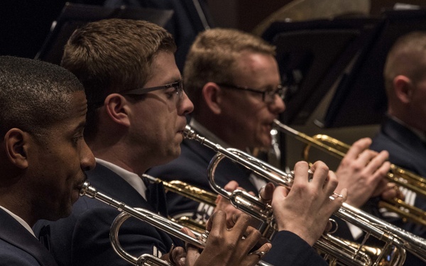 Four military musicians in dress uniforms are playing trumpets during a formal concert or performance. The musicians are seated in a row, all focused on playing their instruments, with music stands visible in the background. The image captures a trumpet section of what appears to be a military band.