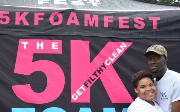 Two people pose together in front of a large promotional banner for 'The 5K Foam Fest' event. A man and woman are embracing and smiling at the camera, both wearing white t-shirts and appearing to be participants in the race. The event's hashtag '#5KFOAMFEST' and tagline 'GET FILTHY CLEAN' are prominently displayed on the banner.