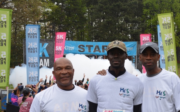 Three men pose together at the starting line of a 5K Foam Fest race, wearing white t-shirts with race bibs numbered 03425, 10578, and 29300. Behind them, foam or smoke is being released at the START line while other participants celebrate in the background. The event is well-branded with multiple '5K Foam Fest' banners and flags surrounding the area.