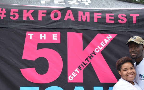 Two people pose together in front of a large promotional banner for 'The 5K Foam Fest' event. A man and woman are embracing and smiling for the photo, both wearing white t-shirts. The woman has a race bib number (0351) visible, indicating they are participants in the event.