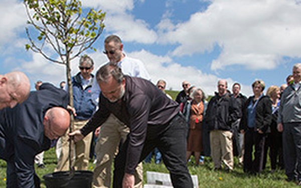 A group of people are gathered outdoors on a grassy hillside for what appears to be a ceremonial tree planting event. Three men in the foreground are actively working to plant a young tree from a black container pot, while a crowd of approximately 20-30 observers watches from behind. The setting appears to be a formal or commemorative occasion based on the organized gathering and attire of the attendees.