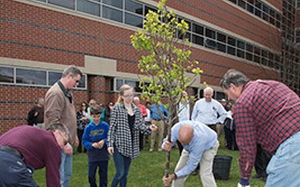 A group of people are participating in a ceremonial tree planting on the grounds of a brick institutional building. Several individuals are actively shoveling soil around a newly planted young tree while others observe. The event appears organized and community-oriented, possibly a memorial or dedication ceremony.