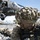 A U.S. Army soldier wearing MultiCam camouflage and a ghillie-covered helmet is in a prone or low-crouch position holding an assault rifle, with a UH-60 Black Hawk helicopter visible in the background. A second soldier is visible lying on the ground to the left. The scene appears to depict a tactical training exercise or combat operation, likely an air assault or helicopter insertion scenario.