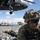 A U.S. Army soldier in full combat gear is prone on rocky terrain, holding a rifle in a security posture beneath a hovering UH-60 Black Hawk helicopter. Multiple other soldiers are visible in the background in similar prone positions, suggesting a tactical air assault or helicopter insertion exercise. The scene appears to be a military training operation conducted in a volcanic or rocky landscape.