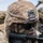 A soldier wearing MultiCam camouflage uniform and helmet is in a prone or low firing position, aiming a rifle. The name tape on the helmet reads 'ZHANG'. The soldier is equipped with tactical ballistic eyewear and a night vision mount bracket on the helmet.