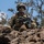 A soldier in full combat gear is positioned on a rocky outcrop, holding an assault rifle and appearing to communicate or issue commands. The soldier is equipped with multicam camouflage uniform, tactical vest, ballistic helmet, and protective eyewear. The setting appears to be a rocky, arid terrain with sparse vegetation under a clear blue sky.