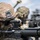 A soldier identified by the name tape 'ZHANG' on their helmet is aiming down the sights of an M4/M16-style assault rifle equipped with an optical scope. The soldier is wearing MultiCam/OCP camouflage pattern uniform and helmet with a night vision goggle mount bracket. Additional soldiers and what appears to be military vehicles are visible in the blurred background.