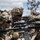 The image shows multiple soldiers in full combat gear taking up defensive firing positions behind rocky terrain. They are equipped with assault rifles featuring optical scopes and tactical accessories, wearing camouflage uniforms and helmets with foliage concealment. The setting appears to be an outdoor training exercise or tactical operation in a rocky, arid environment.