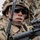 A close-up photograph of a soldier in full combat gear, wearing OCP (Operational Camouflage Pattern) uniform and helmet with camouflage netting. The soldier is holding what appears to be an M4 carbine rifle equipped with an ACOG (Advanced Combat Optical Gunsight) scope. The image captures the soldier in an alert, ready posture suggesting a training exercise or operational deployment.