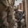A soldier in full combat gear navigates through a narrow passage between wooden barriers or crates. The soldier is wearing MultiCam camouflage, a helmet with camouflage netting, body armor, and carrying a rifle with an underbarrel grenade launcher attachment. The image appears to be taken during a military training exercise or operational movement.