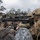 A soldier in full multicam camouflage gear is positioned prone among volcanic rocks, aiming a precision sniper rifle equipped with a high-powered scope and bipod. The setting appears to be a rocky, lava field terrain with sparse vegetation and an overcast sky. The image captures a military training or operational scenario with professional-grade equipment.