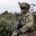 A U.S. Army soldier in full combat gear is photographed in a rocky, vegetated outdoor environment. The soldier wears MultiCam (OCP) camouflage, a helmet with camouflage netting and mounted equipment, and carries a rifle and tactical backpack. A unit patch consistent with the 25th Infantry Division ('Tropic Lightning') is visible on the left shoulder.