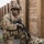 A U.S. Army soldier in full combat gear is positioned against a wooden wall structure, appearing to be in a tactical training exercise or operational environment. The soldier is equipped with multicam camouflage uniform, helmet with foliage, body armor, and is holding an assault rifle. The setting appears to be a military training facility with a constructed wooden barrier corridor.