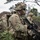 A U.S. Army soldier identified by the name tape 'Vasquez' is shown in full combat gear during what appears to be a field training exercise. The soldier is equipped with an M4-style carbine, a rocket launcher (AT4 or similar) strapped to their backpack, and has camouflage face paint applied. A second soldier is visible in the background in a similar operational posture.