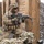 A soldier in full combat gear is kneeling in a tactical position behind wooden post cover, aiming an assault rifle. The setting appears to be a military training range, identifiable by the constructed wooden barrier lane and gravel/rock ground surface. A second soldier or equipment bundle is partially visible in the foreground.