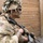 A soldier in full combat gear is positioned against a wooden wall structure, holding an assault rifle in a tactical stance. The soldier is wearing MultiCam/OCP camouflage uniform, a helmet with ghillie-style foliage camouflage, tactical sunglasses, and face paint. The image appears to be taken during a military training exercise, evidenced by the 'SAFETY' label visible on equipment.
