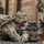 Military personnel in full combat gear are conducting what appears to be a tactical training exercise near a wooden structure. The central soldier is in a prone position aiming a rifle with an optical sight, while other soldiers are visible in the background and foreground. All personnel are wearing multicam/OCP camouflage uniforms with full kit including helmets, body armor, and tactical backpacks.