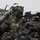 A heavily equipped soldier in MultiCam/OCP camouflage is positioned behind volcanic rock terrain, aiming an M4 carbine rifle with an optic sight. The soldier carries multiple pieces of kit including what appears to be an AT4 or similar rocket launcher on their back, along with a full tactical load. The setting and equipment suggest a U.S. Army training exercise or operational deployment.